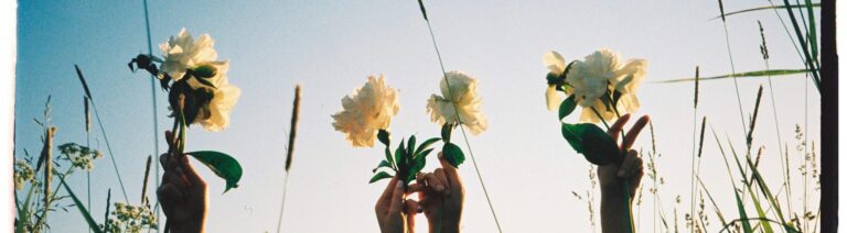 Two women raise white flowers in a sunny field, capturing summer joy and freedom.